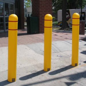 Three yellow bollards with red bands, similar to the Vestil Plastic Bollard Post Cover Red Tape 52X5, stand on a concrete sidewalk by a building. A tree, trash bin, and parked vehicle fill the busy urban scene.