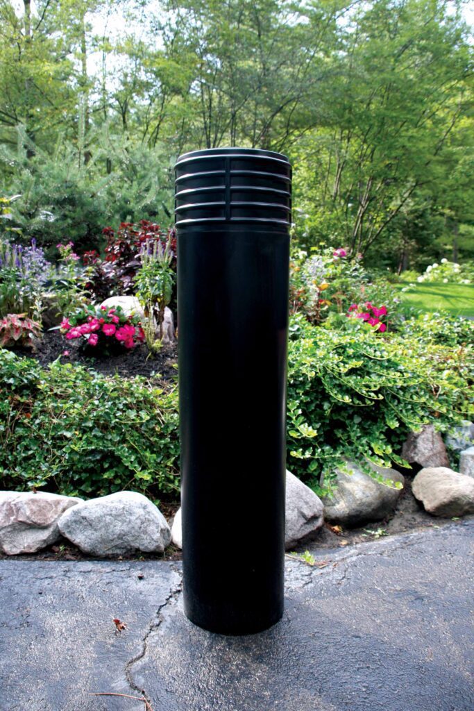 BPC-DC-B_A.jpg A Vestil Cinco-Black Bollard Cover 52 In stands on a paved surface with stone-bordered landscaping. Lush greenery and colorful flowers are abundant in the background under a canopy of trees.