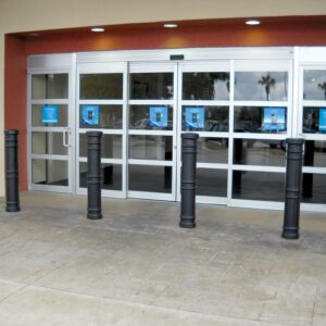 Automatic sliding glass doors with reflective panels are set against a soft yellow and red wall, protected by five black bollards each with a 52-inch Vestil Metro-Black Bollard Cover, on patterned stone pavement. Blue-background signs adorn each door panel.