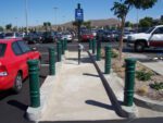 The parking lots cart return area is lined with Vestil Metro-Green Bollard Cover 52 In posts along a concrete path. With parked cars flanking, shady trees, a blue cart return sign, and distant hills under clear skies complete the scene.