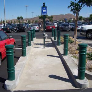 The parking lots cart return area is lined with Vestil Metro-Green Bollard Cover 52 In posts along a concrete path. With parked cars flanking, shady trees, a blue cart return sign, and distant hills under clear skies complete the scene.