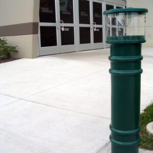 A Vestil Metro-Green Bollard Cover-Ac Light 57 In, featuring a transparent section, stands on a concrete sidewalk near glass-door building entrance, with small plants visible in the background.