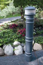A tall, dark outdoor lamp post with a 57-inch Vestil Metro-Gray Bollard Cover-Ac Light adorns a stone pathway, enhanced by rocks and red & white flowers. Green foliage and a driveway add charm to the garden scene in the background.