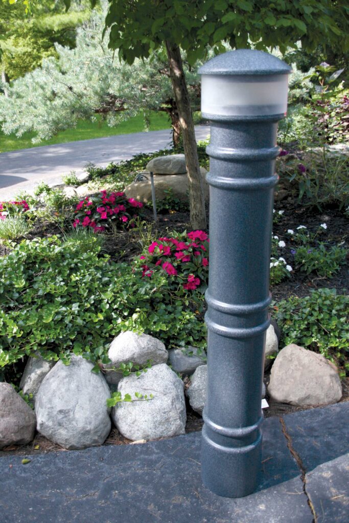 A tall, dark outdoor lamp post with a 57-inch Vestil Metro-Gray Bollard Cover-Ac Light adorns a stone pathway, enhanced by rocks and red & white flowers. Green foliage and a driveway add charm to the garden scene in the background.