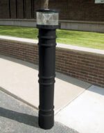 A paved pathway beside a brick wall features a black bollard with a transparent, dome-shaped light resembling the 57-inch Vestil Metro-Black Bollard Cover W/Light. Grass and a building can be seen in the background.