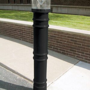 A paved pathway beside a brick wall features a black bollard with a transparent, dome-shaped light resembling the 57-inch Vestil Metro-Black Bollard Cover W/Light. Grass and a building can be seen in the background.