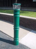 A tall, green cigarette disposal bin stands on a sidewalk next to a brick wall. The design resembles a Vestil Metro-Green Bollard Cover W/Light 57 In, with a clear section at the top for viewing contents. Pavement and grass are visible in the background.