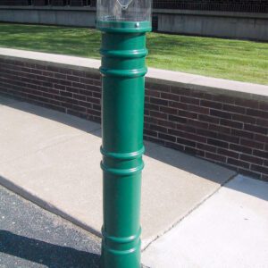 A tall, green cigarette disposal bin stands on a sidewalk next to a brick wall. The design resembles a Vestil Metro-Green Bollard Cover W/Light 57 In, with a clear section at the top for viewing contents. Pavement and grass are visible in the background.