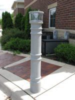 A Vestil Metro-Gray Bollard Cover W/Light 57 In stands near a brick building with green shrubs on a sidewalk. The cylindrical light has a clear top, surrounded by paved and concrete surfaces under a clear sky.