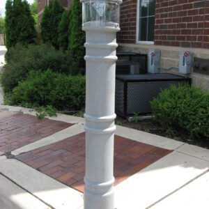 A Vestil Metro-Gray Bollard Cover W/Light 57 In stands near a brick building with green shrubs on a sidewalk. The cylindrical light has a clear top, surrounded by paved and concrete surfaces under a clear sky.