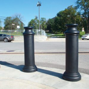 Two large, black cylindrical bollards on a sidewalk by a road with cars in the background. Trees, grass, and fencing under a clear blue sky complete the scene, enhanced by the sleek Vestil Pawn-Black Bollard Cover 49 In.