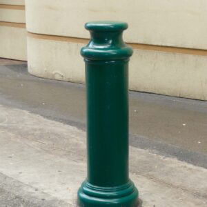 A Vestil Pawn-Green Bollard Cover 49 In stands on a concrete surface near a building, its tall, green cylindrical shape with rounded top enhancing modernity and safety along the sidewalk against the smooth wall backdrop.