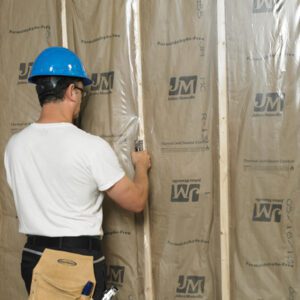 A construction worker in a blue hard hat and white shirt expertly installs wall insulation, securing the brown material to wooden beams with a staple gun. His tool belt includes MH-USA 12 x 100 - 4 Mil Clear Poly Sheeting (1/Case) for added precision and efficiency.