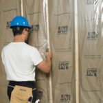 A construction worker in a white shirt and blue helmet staples MH-USA 6 x 100 - 6 Mil Clear Poly Sheeting (1/Case) to wooden wall studs. With his tool belt ready, he focuses on securing the material, complete with text and logos, ensuring optimal insulation.