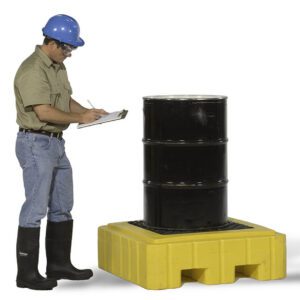A person in a blue hard hat, safety goggles, tan shirt, jeans, and black boots writes on a clipboard next to a black barrel on a yellow Vestil Ultra Spill Pallet.