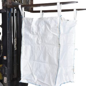A large white bulk bag, resembling the Vestil Fabric Hopper 3.3K 50 Cu Ft, hangs from the forklift forks. With dimensions of 36 X 36 X 55 and reinforced lifting loops, it’s designed for heavy-duty transport. The forklift is partially visible on the left side.