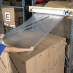 Someone stretches clear plastic wrap from a mounted dispenser over a stack of cardboard boxes on a shelf, with 60 x 60 Goodwrappers® Top Sheeting (4 Rolls/Case) nearby. Other boxes and packing materials are scattered throughout the warehouse, creating an organized yet bustling storage area.