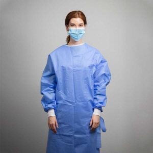 A person with red hair stands facing forward, wearing a blue medical gown and a blue face mask. The background is a plain, light-colored wall.