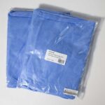 Two packages of folded blue scrub tops are sealed in transparent plastic. Each package has a white label displaying text. The fabric appears smooth and is neatly packed, with the tops intended for medical or professional use.