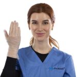 A woman in a blue medical scrub top holds up her right hand, wearing a clear disposable glove. She is smiling and facing the camera against a plain white background.