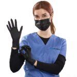 A woman in blue medical scrubs and a black face mask is putting on black gloves. She has red hair tied back and is standing in front of a white background. She raises one hand while adjusting the glove with the other.