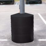 A Vestil Light Pole Base Protector Black surrounds the gray lamppost base in a parking lot, forming a cylindrical barrier from three stacked segments, with parking spaces and partial car views in the background.
