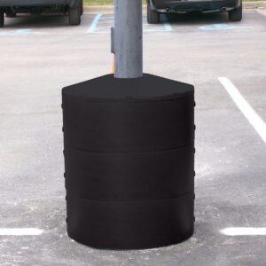 A Vestil Light Pole Base Protector Black surrounds the gray lamppost base in a parking lot, forming a cylindrical barrier from three stacked segments, with parking spaces and partial car views in the background.