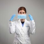 A person with red hair is in a white lab coat and blue gloves, holding a blue surgical mask in front of their face, surrounded by custom laboratory casework. The background is plain gray, enhancing the focus on high-performance lab sinks as part of complete laboratory solutions.