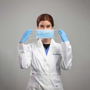 A person with red hair is in a white lab coat and blue gloves, holding a blue surgical mask in front of their face, surrounded by custom laboratory casework. The background is plain gray, enhancing the focus on high-performance lab sinks as part of complete laboratory solutions.