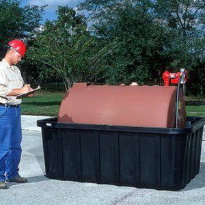 A person in a red hard hat and safety glasses is taking notes on a clipboard next to a large brown cylindrical container in a black 550 lb Vestil Fuel Tank Containment with Drain. Trees can be seen in the background, enhancing the tranquil atmosphere.