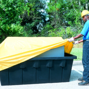 A person wearing a hard hat and gloves adjusts a Vestil Containment Sump Otc-275 Pull Over Cover on a large black container outdoors, with trees and grass enhancing the natural backdrop.