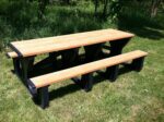 A Vestil Picnic Table with a cedar top and two benches, featuring the Poly Walk Thru 2896 BK Leg design, sits on the grass. Contrasting black supports amid trees suggest a sunny day—a perfect spot to enjoy nature.