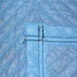 Close-up of a quilted blue fabric corner, similar to Vestil General Duty Moving Pads Cotton 1 Pack. Black stitches intersect at right angles, with textured cotton material and diagonal seams creating a braided pattern.