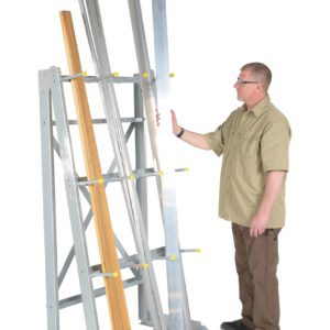 A man in a khaki shirt and brown pants carefully places long metal beams onto the Vestil Vertical Floor Mounted Rack W/ 2000 Lb, which also holds wooden beams, set against a plain white background.