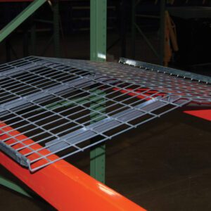 The orange and green warehouse pallet rack holds a Vestil Pallet Rack-Crown Wire Deck 58 X 47.6 In. Its slanted wire mesh, detachable design, and versatility are emphasized. The warehouse floor serves as a backdrop.