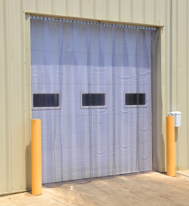 A beige industrial building showcases a Vestil Vinyl Strip Door 0.08 In Full 108 X 48 with clear plastic and three rectangular windows. Two yellow bollards protect the entrance on the concrete, ensuring safety and functionality.