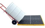 A white barrel rests on a red hand truck, close to a Vestil Ultra Spill Deck Four Drum Flexible. The black and yellow spill platform features a grid pattern to prevent spills from reaching the ground.
