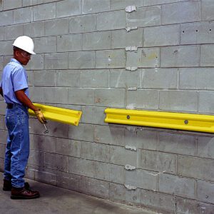Wearing a hard hat and safety glasses, a person installs a Vestil Polyethylene Wall Protector 48 In on a gray cinder block wall using a tool, while another yellow bumper rail is already secured beside it.