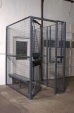 A Vestil Driver/Trucker Access Cage 3 Sided 4X4, featuring a sturdy metal door and wire mesh sides enclosing a bench, is tucked in the corner of a room with concrete floors and pristine white walls.