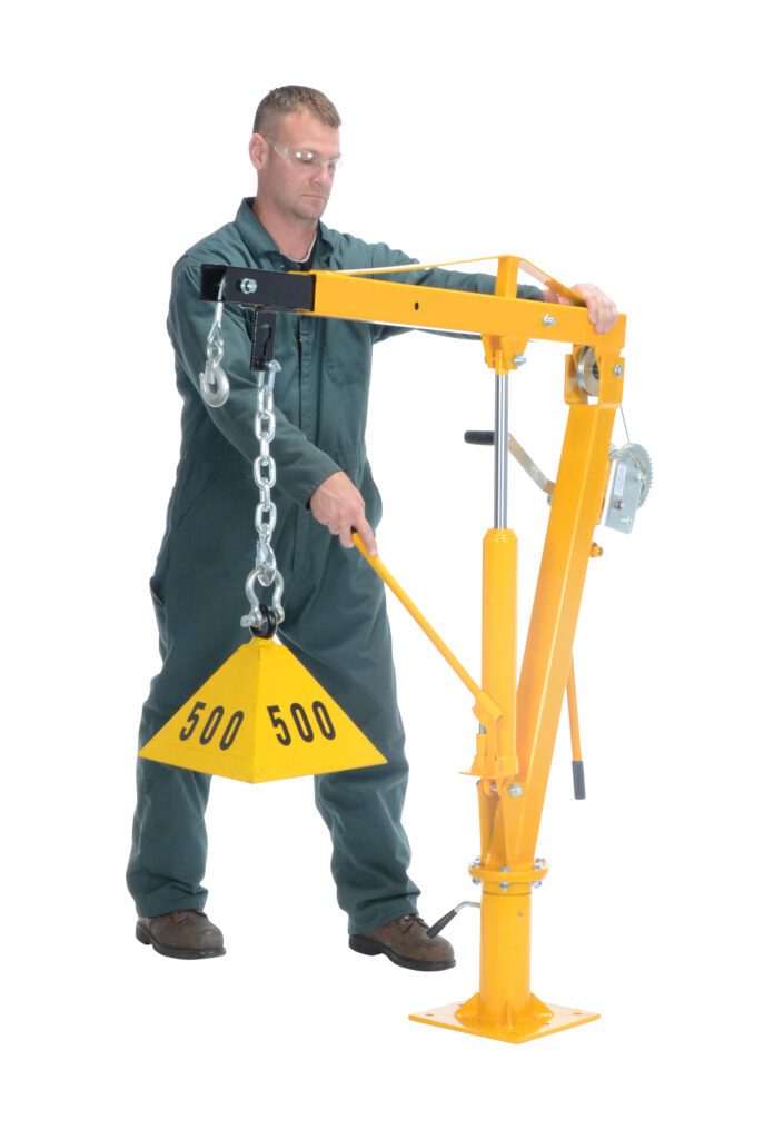 In a green jumpsuit, a person operates a yellow Vestil Winch Operated Truck Jib Crane 1K Extend, handling the triangular 500 weight with precision. The device has a manual crank and is set against a plain white backdrop.