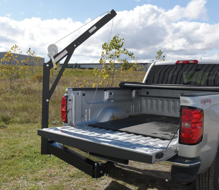 A silver pickup with its tailgate open is parked on a grassy field, showcasing a Vestil Hitch-Mounted Truck Jib Crane with a winch over the tailgate. The peaceful setting includes small trees and a partly cloudy sky.