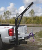 A pickup truck with an open tailgate features the Vestil Hitch-Mounted Truck Jib Crane, which uses a pulley, rope, and purple harness to lift objects. A grassy field and trees are visible in the background under a partly cloudy sky.