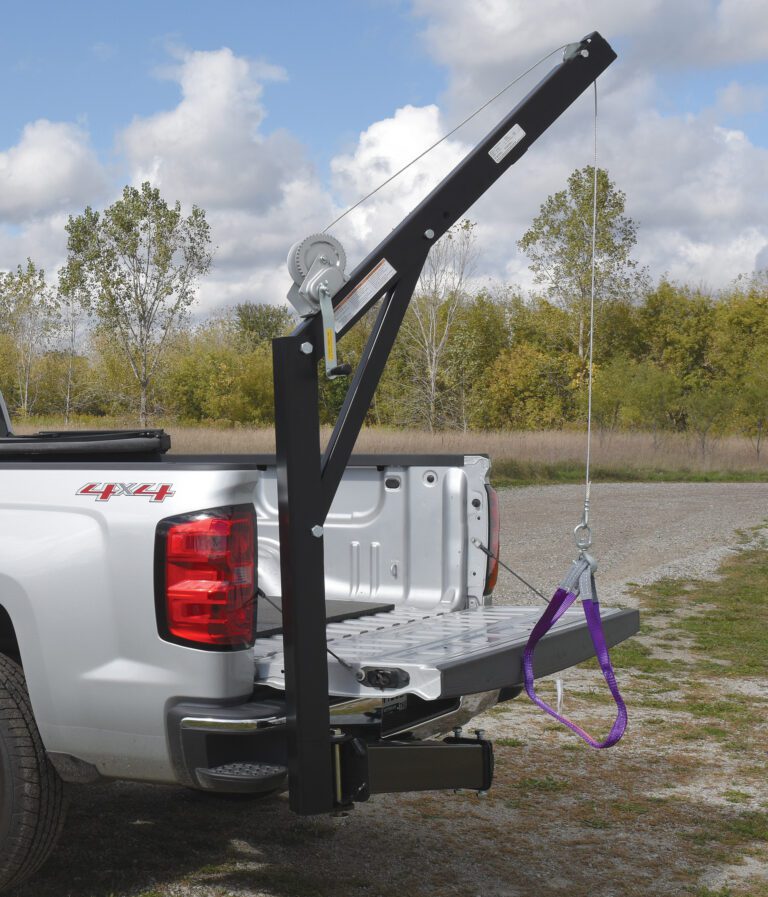 A pickup truck with an open tailgate features the Vestil Hitch-Mounted Truck Jib Crane, which uses a pulley, rope, and purple harness to lift objects. A grassy field and trees are visible in the background under a partly cloudy sky.