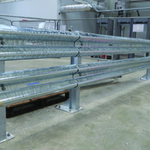 A metal safety barrier with yellow ends is installed indoors on a concrete floor. The barrier features two horizontal rails and vertical supports. In the background, there are metal gates and construction equipment.