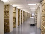 An archive room with bright lighting features Aurora Quik-Lok Shelving, adjustable heavy-duty steel units with 350 lbs capacity. Stacked brown boxes line the shelves, enhancing a clean atmosphere on a gray floor and white ceiling, while a large gray cart stands at the center.