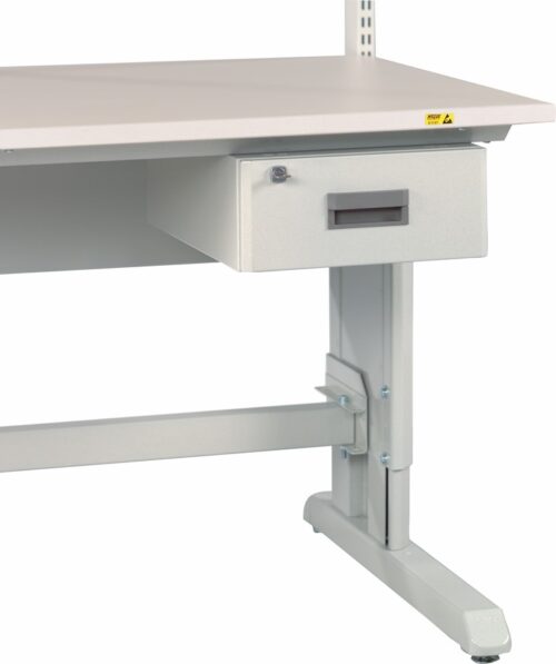 A gray metal workbench with an attached drawer beneath the tabletop. The bench has an adjustable support frame, visible legs, and a small yellow warning label on the corner of the tabletop.