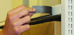 A person applies a magnetic label holder to a metal shelf edge. The holder is curved and dark in color. Below the shelf, a cardboard box is partially visible. The background is out of focus.