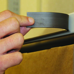 A person applies a magnetic label holder to a metal shelf edge. The holder is curved and dark in color. Below the shelf, a cardboard box is partially visible. The background is out of focus.
