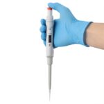 A hand wearing a blue glove holds a white adjustable pipette with precision, its digital display showing measurement settings. Pointed downward, it suggests readiness for use in a laboratory setting, emphasizing the importance of pipette calibration for accurate results.