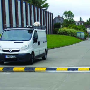 A white van with a ladder on top and license plate YH57 CUC is parked on a paved road near a speed bump. It is adjacent to a building with striped walls, and a row of houses and greenery is visible in the background.
